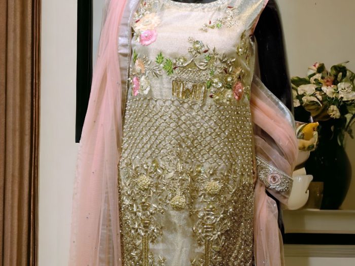 A pink-gold hand-embellished dress crafted from organza zari fabric, featuring intricate detailing. Paired with raw silk pink trousers and a delicate net dupatta, the outfit exudes elegance and luxury.