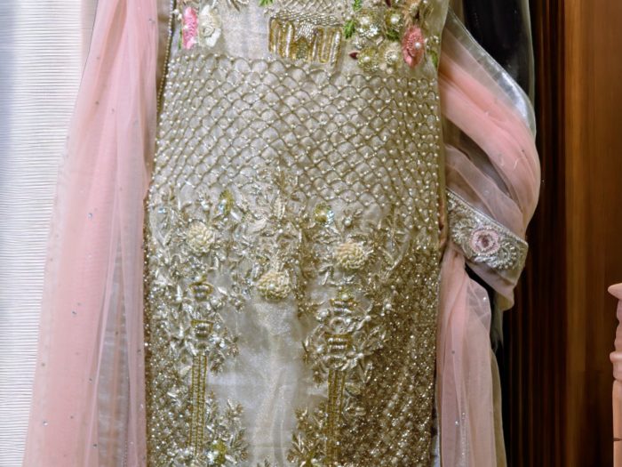 A detailed close-up of the pink-gold organza zari dress, showcasing fine hand embellishments with shimmering accents. The intricate craftsmanship highlights the rich textures and luxurious feel of the fabric.